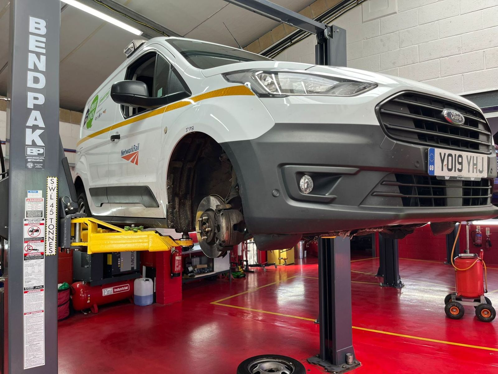 A white fleet van up on a lift in the workshop with its front wheel off